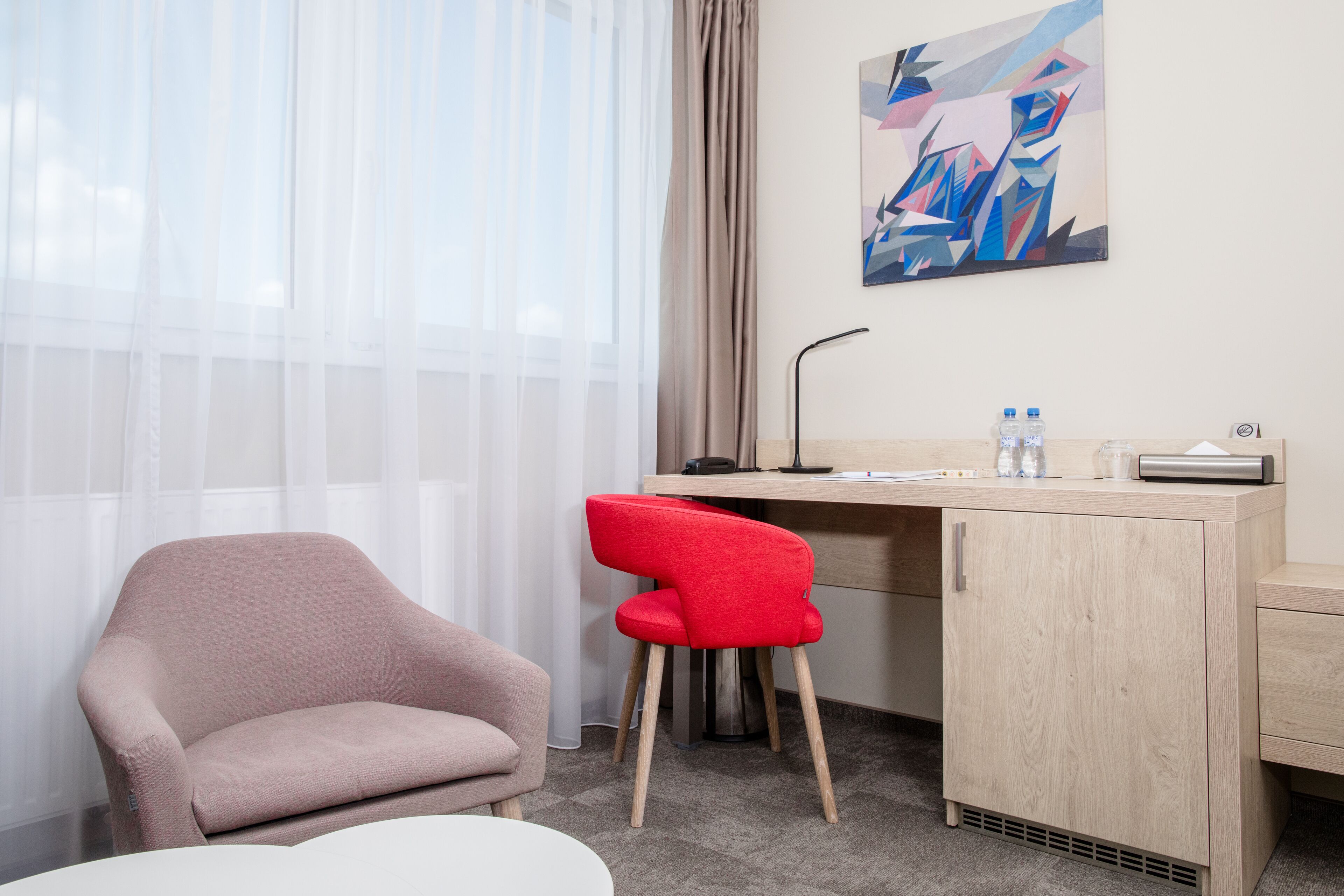 Een hotelkamer met een bureau, een rode stoel, een beige fauteuil en een schilderij aan de muur.
