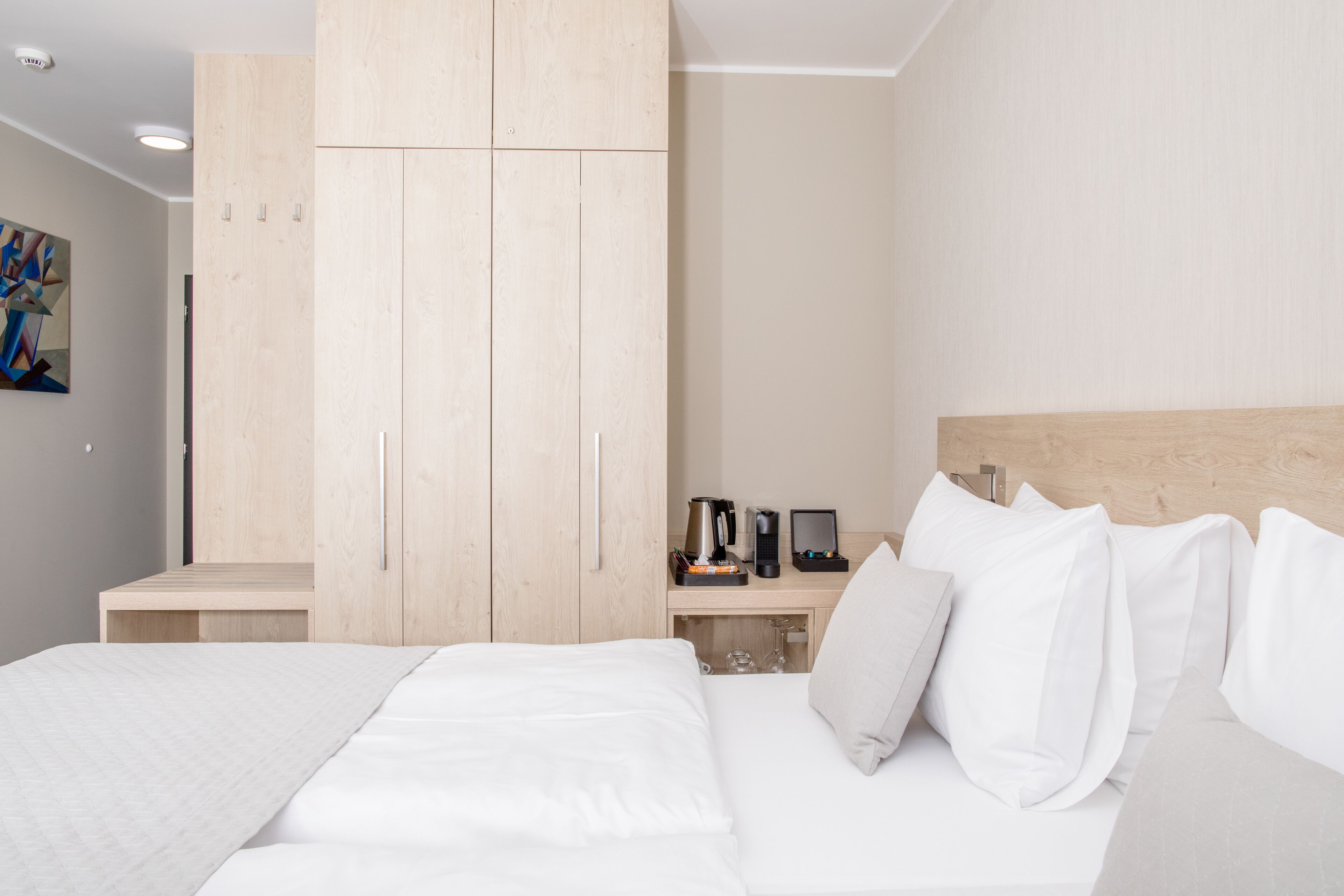 A hotel room with a bed, a wooden wardrobe, a bedside table with a coffee maker, and a painting on the wall.