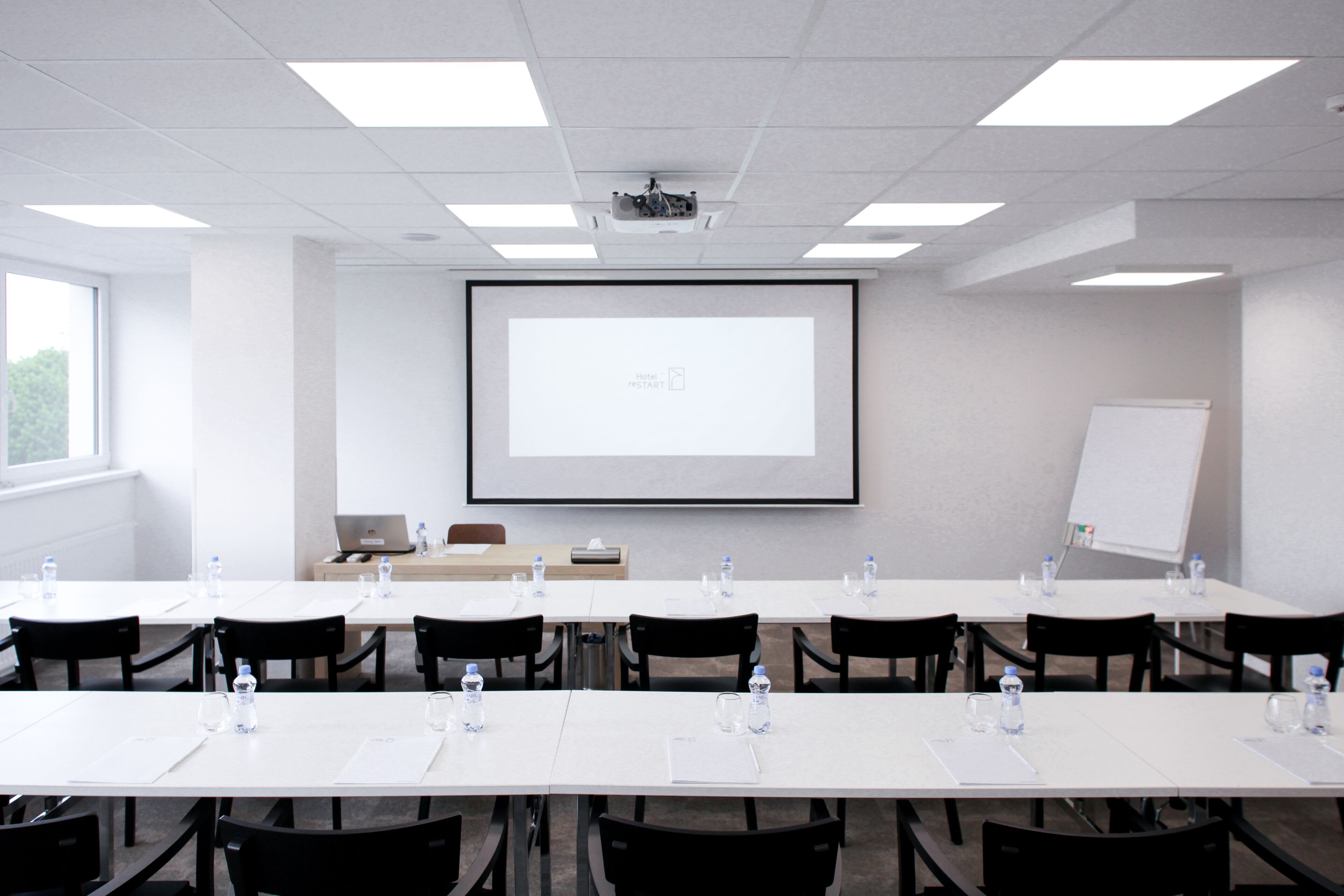 Een vergaderruimte met een groot projectiescherm, witte tafels, zwarte stoelen en een whiteboard.