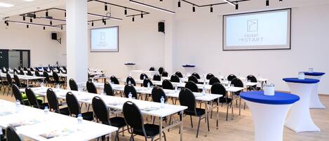 A conference room with rows of tables and chairs, a projector screen, and a large screen displaying 'Hotel restart'.