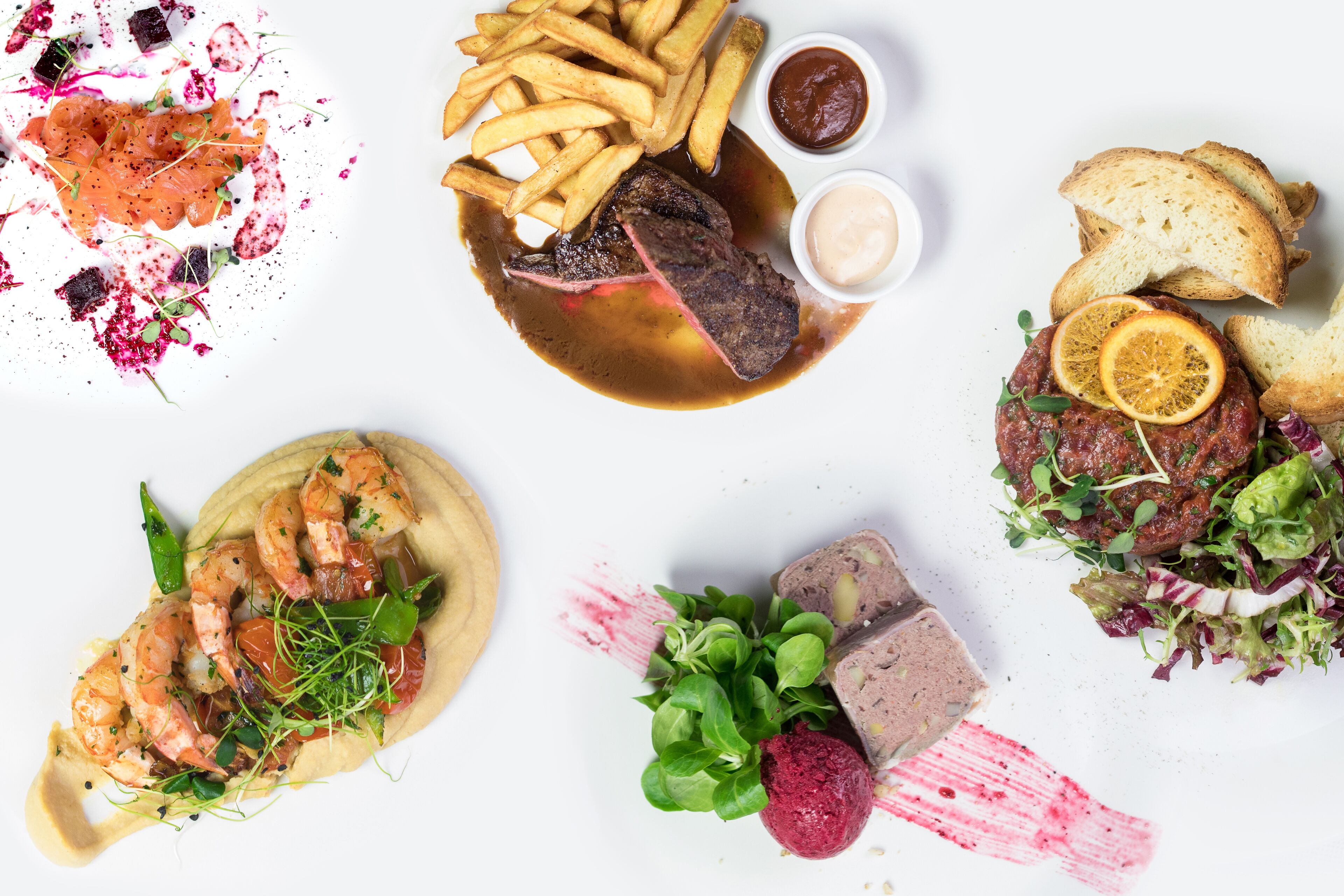 Une variété de plats, notamment des steaks, des frites et des crevettes, disposés sur une surface blanche.