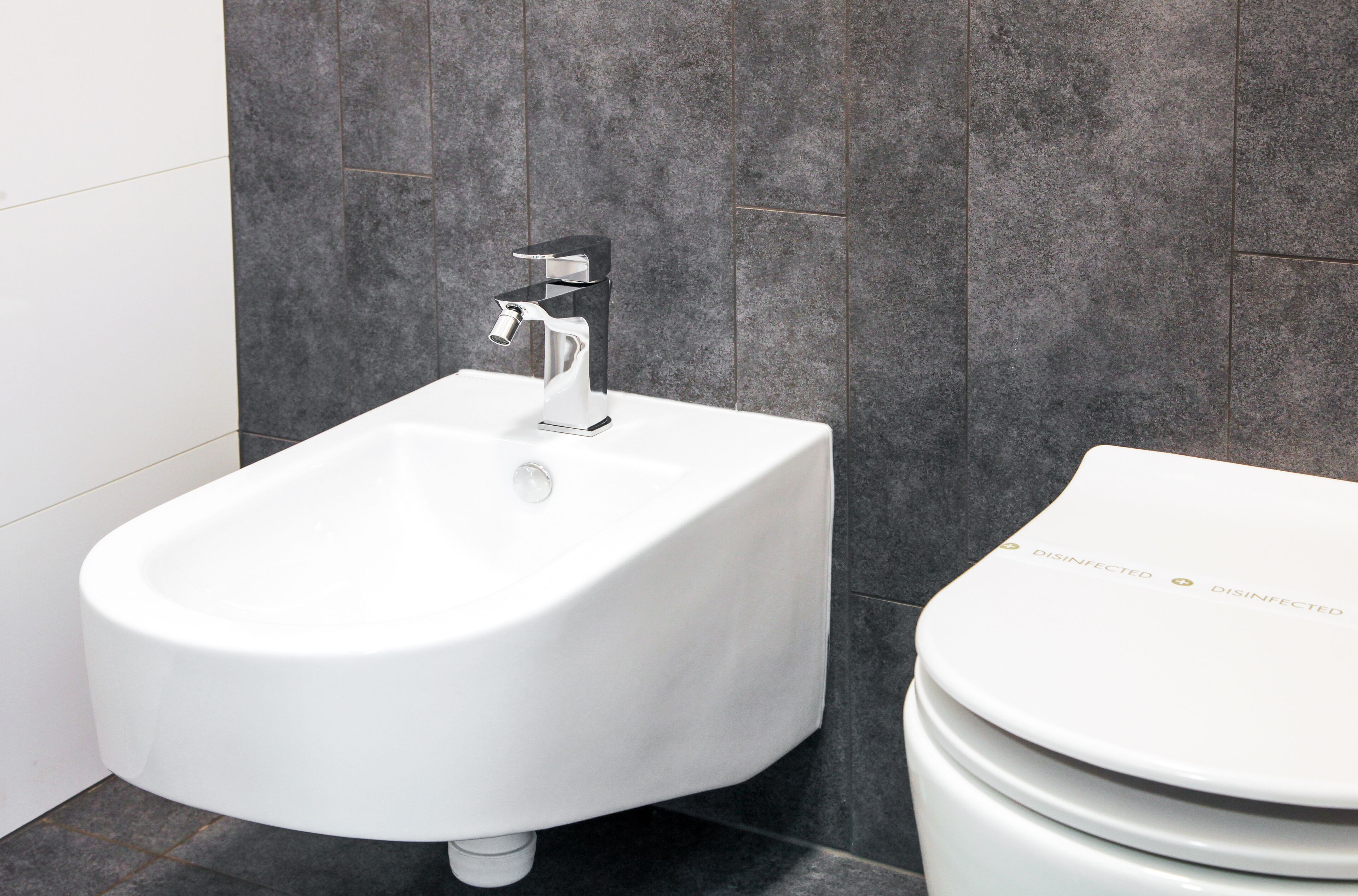 A modern bathroom with a bidet, a toilet, and dark tiled walls.