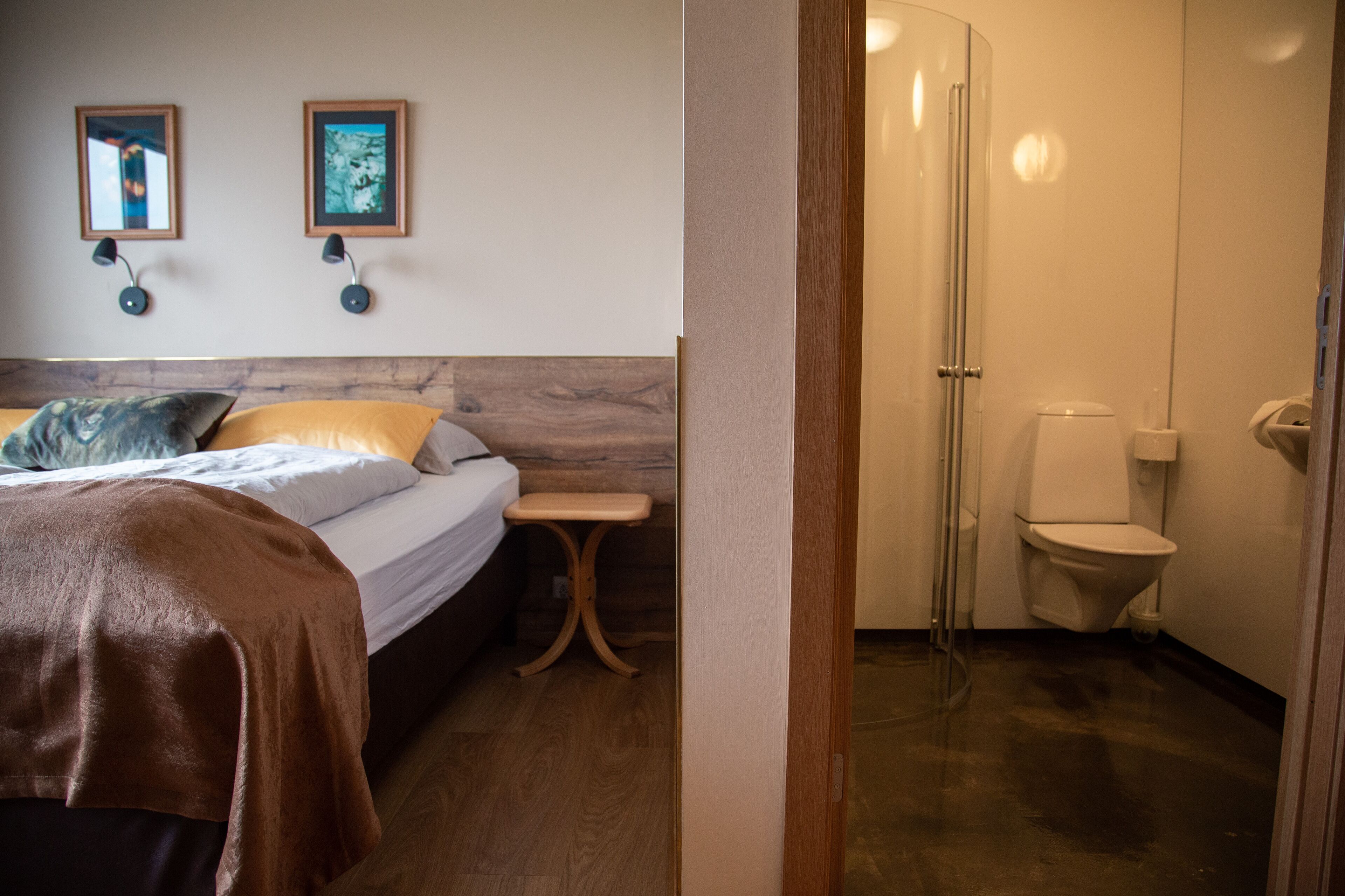 A bedroom with a bed, bedside table, and two wall-mounted lamps. Adjacent is a bathroom with a toilet, shower, and glass door.