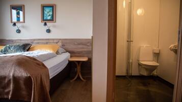 A bedroom with a bed, bedside table, and two wall-mounted lamps. Adjacent is a bathroom with a toilet, shower, and glass door.