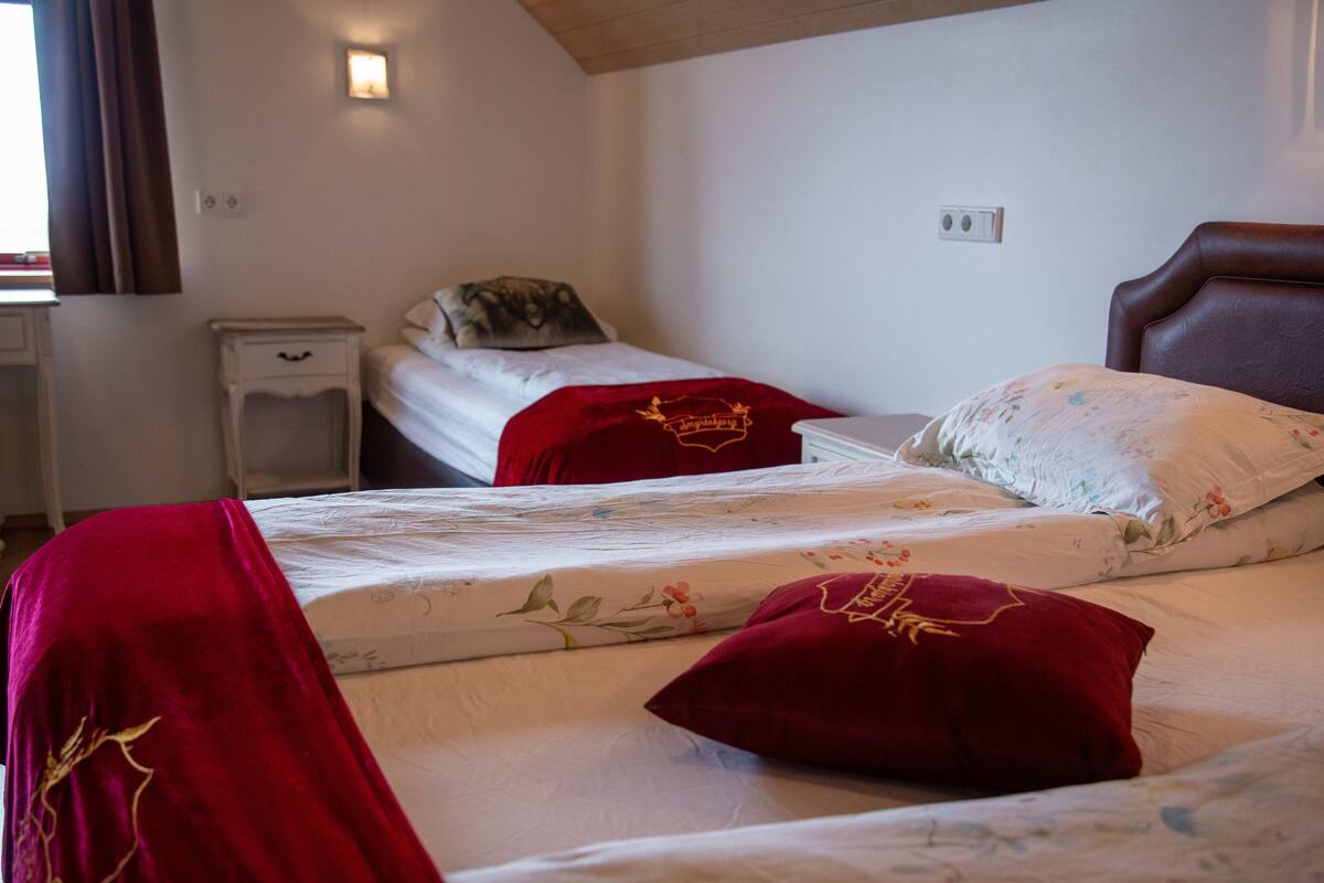 A hotel room with two beds, each with a red blanket and pillow, a nightstand, and a small table with a lamp.