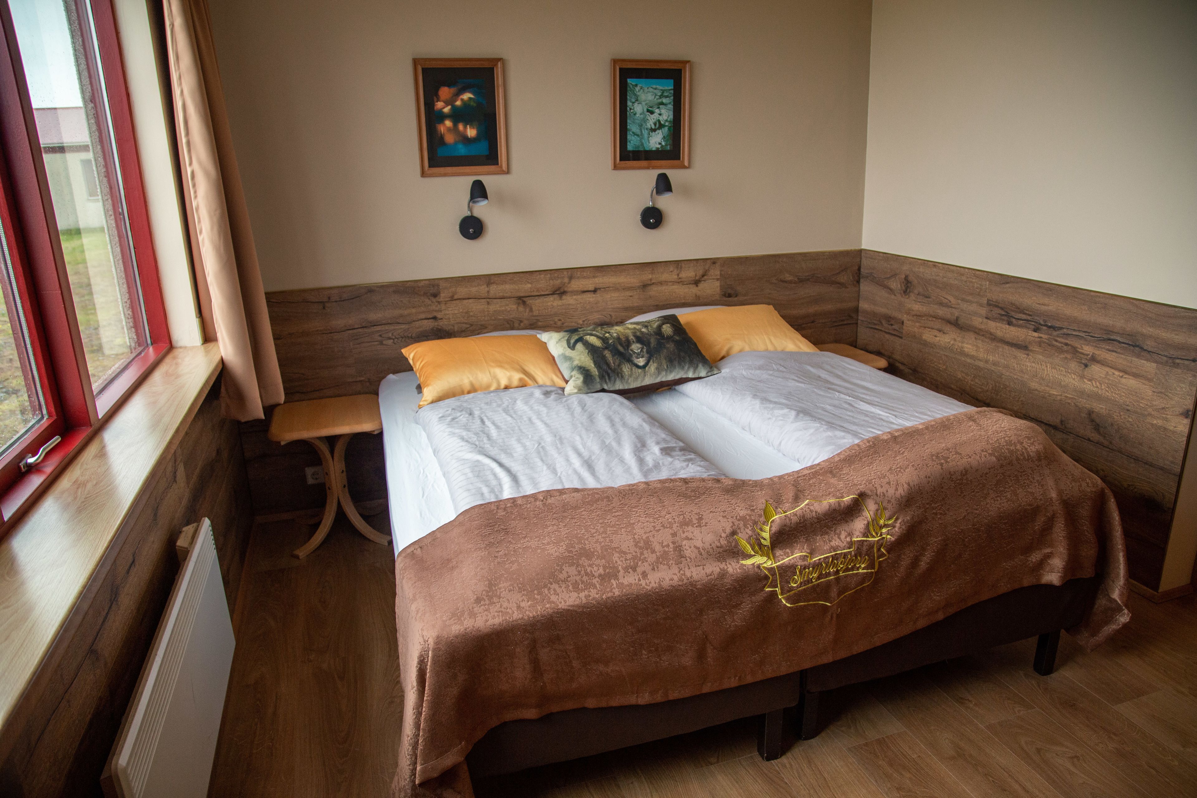 Une chambre à coucher avec une tête de lit en bois, un lit recouvert d’une couverture marron, deux lampes fixées au mur et deux tableaux encadrés.