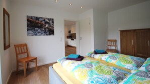 Ein Schlafzimmer mit zwei Betten, einem Holzstuhl, einem Bild an der Wand und Blick zur Küche.