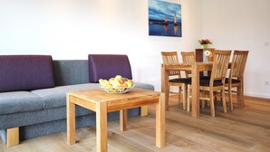 Ein Wohnzimmer mit grauem Sofa, einem Couchtisch mit Obstschale und einem Essbereich mit Holztischen und -stühlen.