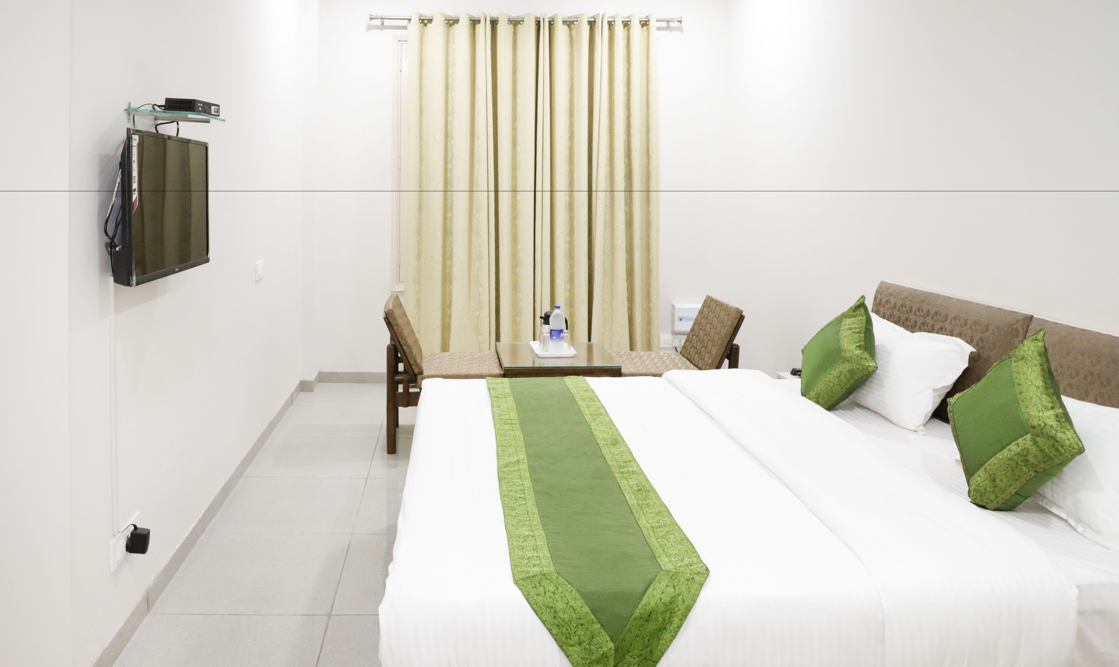 A hotel room with a bed, a TV mounted on the wall, a table with a chair, and a window with curtains.