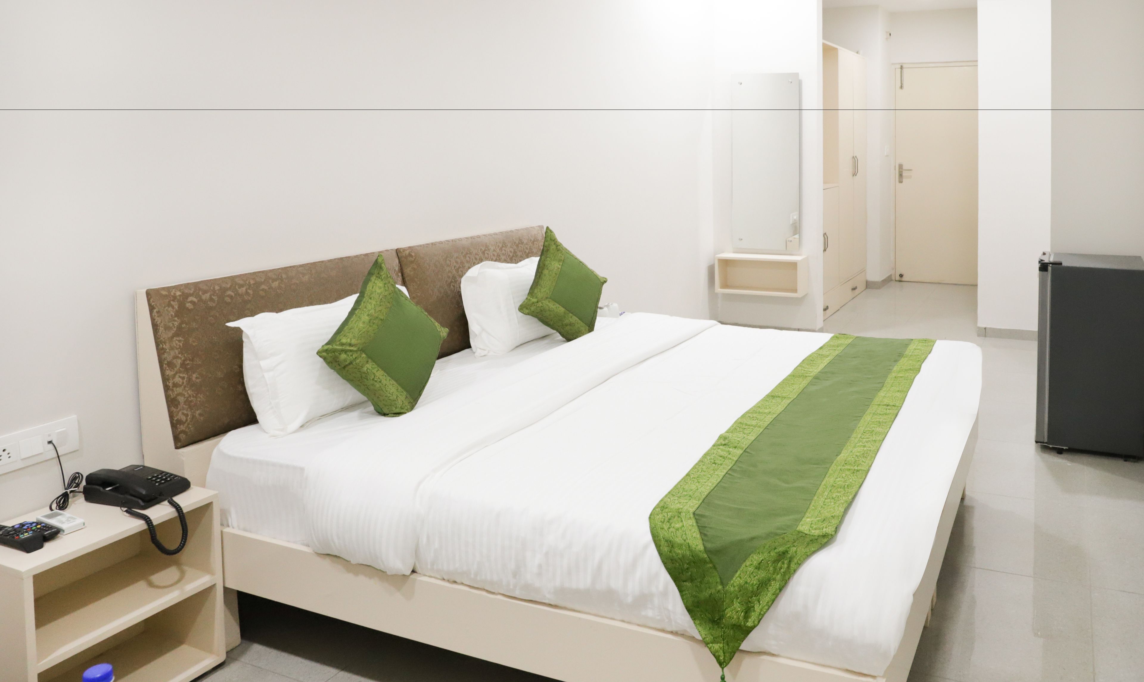 Een netjes opgemaakt bed met witte beddengoed en groene accenten, een telefoon op het nachtkastje, en een klein plankje met een telefoon en oplader.