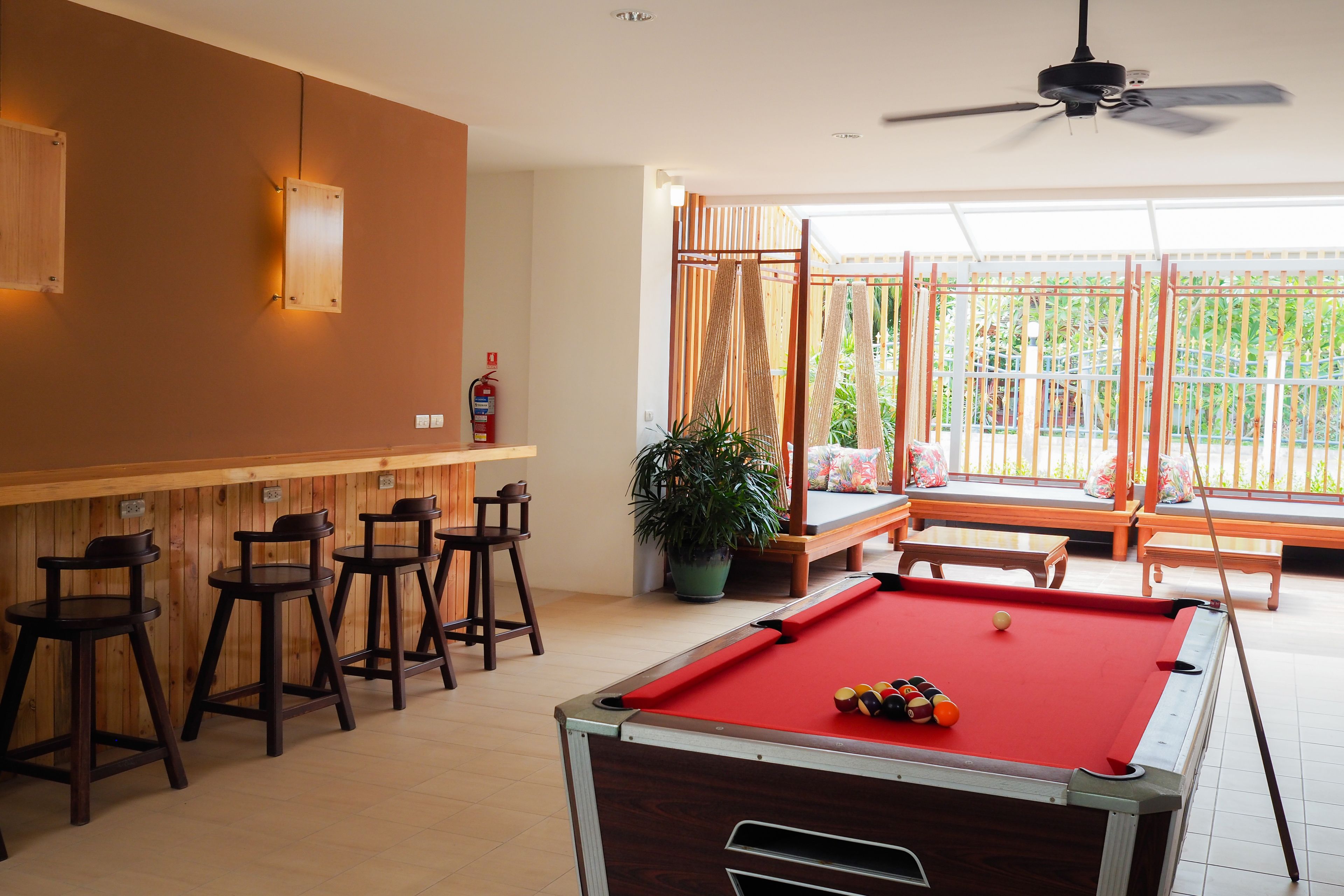 A pool table with a red felt surface, surrounded by wooden bar stools. There is a ceiling fan above and a potted plant near the window.