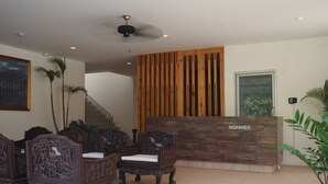 A reception area with a wooden reception desk labeled "NONNEE," several intricately carved wooden chairs, a ceiling fan, and a staircase in the background. - Nonnee (Karon)
