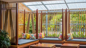 A room with wooden furniture, including a sofa and a table, and a large window with a view of greenery. - Nonnee (Karon)