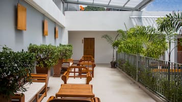 A covered outdoor seating area with wooden tables and chairs, potted plants, and a view of the sky through a glass roof.