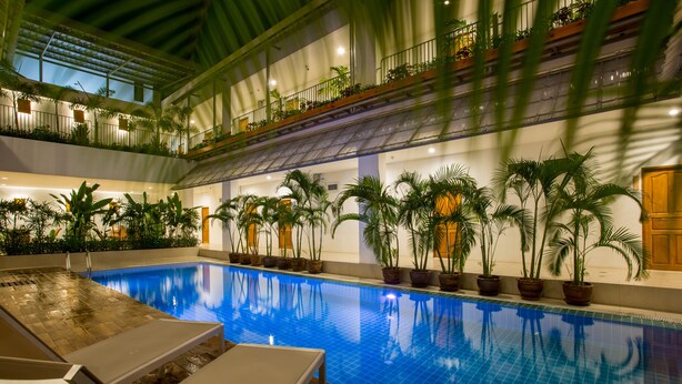 An indoor swimming pool with lounge chairs, potted plants, and a multi-level balcony with railings. - Nonnee (Karon)