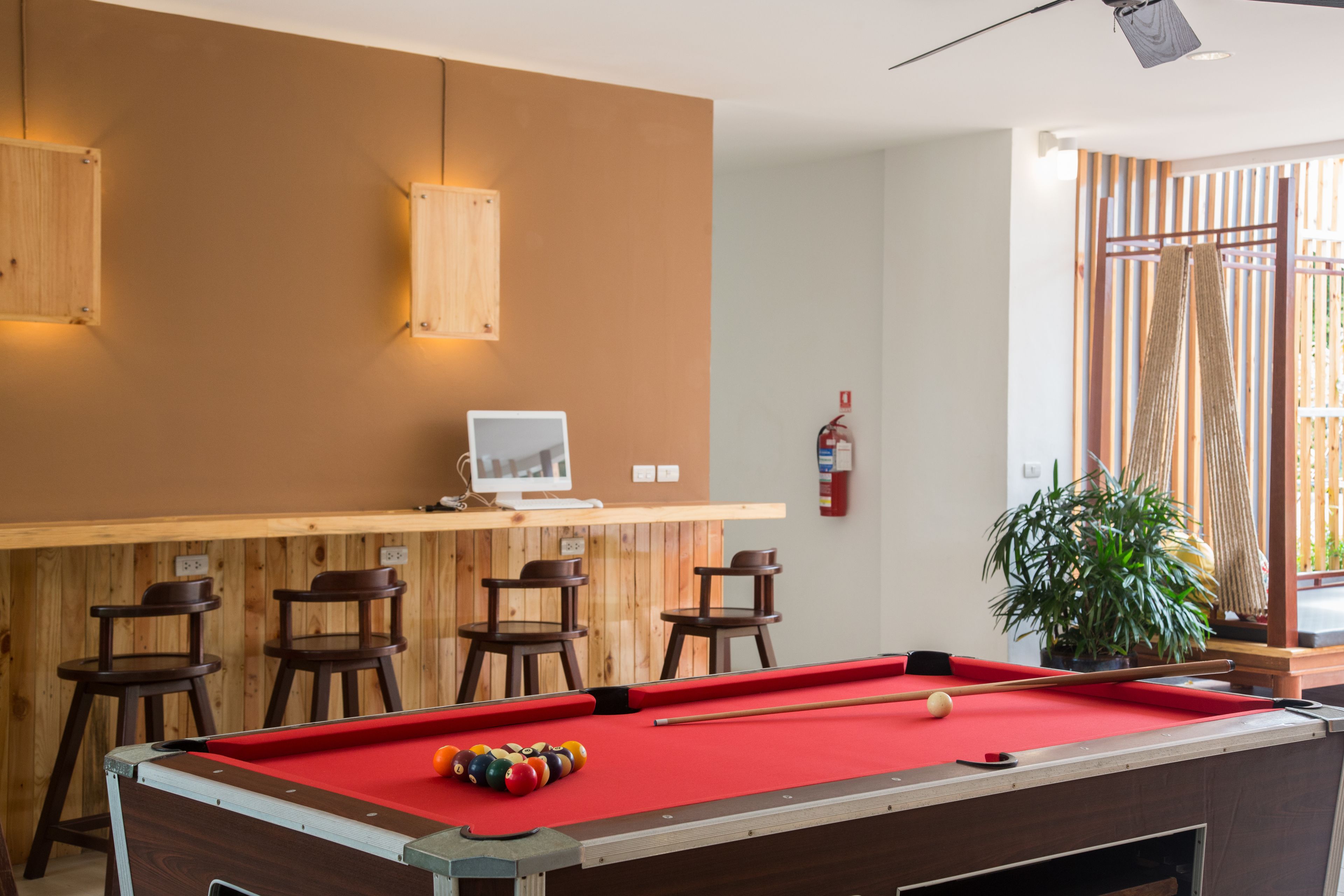 A pool table with a red felt surface, wooden bar stools, a computer monitor on a wooden shelf, and a potted plant.