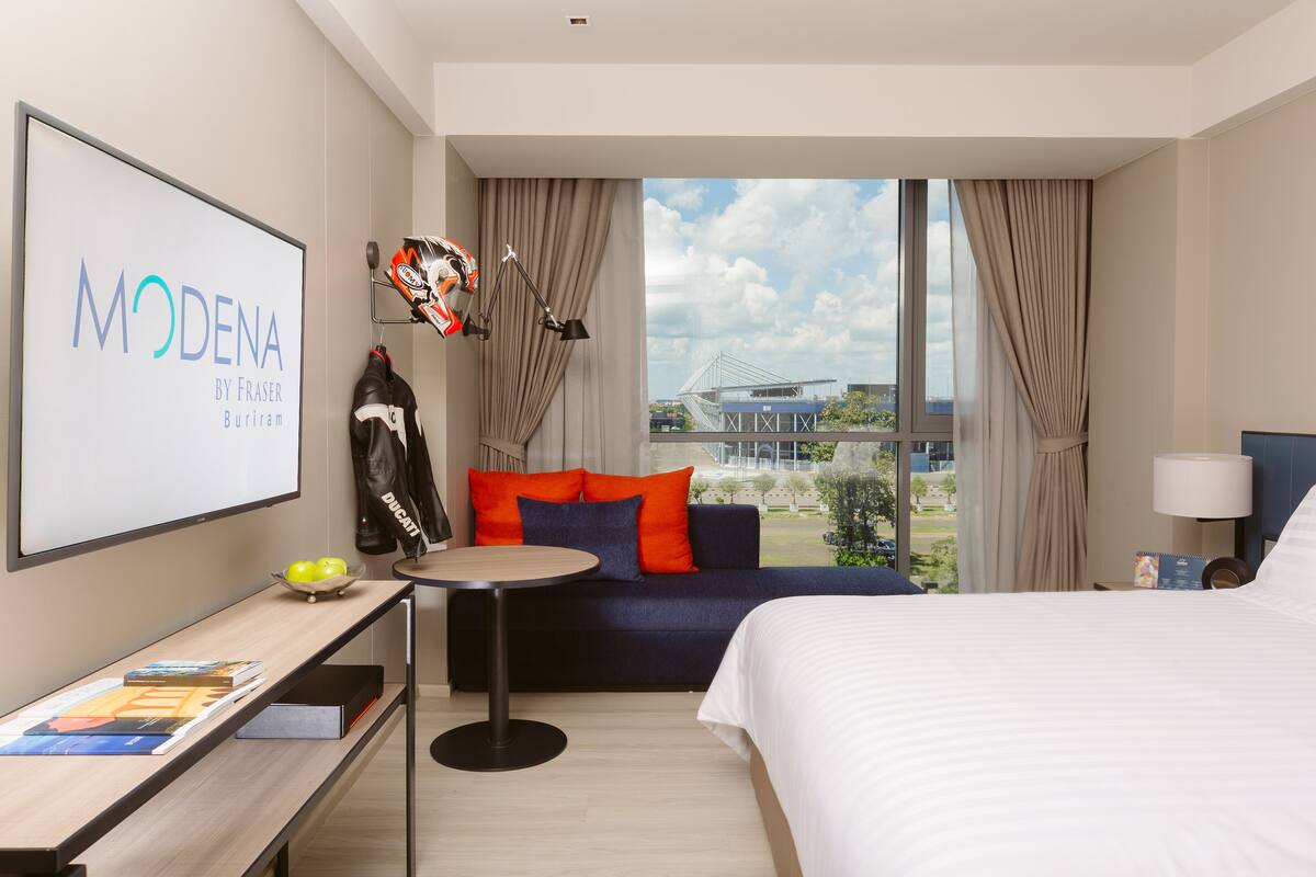 Ein Hotelzimmer mit einem Bett, einem Schreibtisch mit einem Laptop, einer Couch mit bunten Kissen, einem Fenster mit Blick auf ein Gebäude und einem an der Wand befestigten Motorrad.