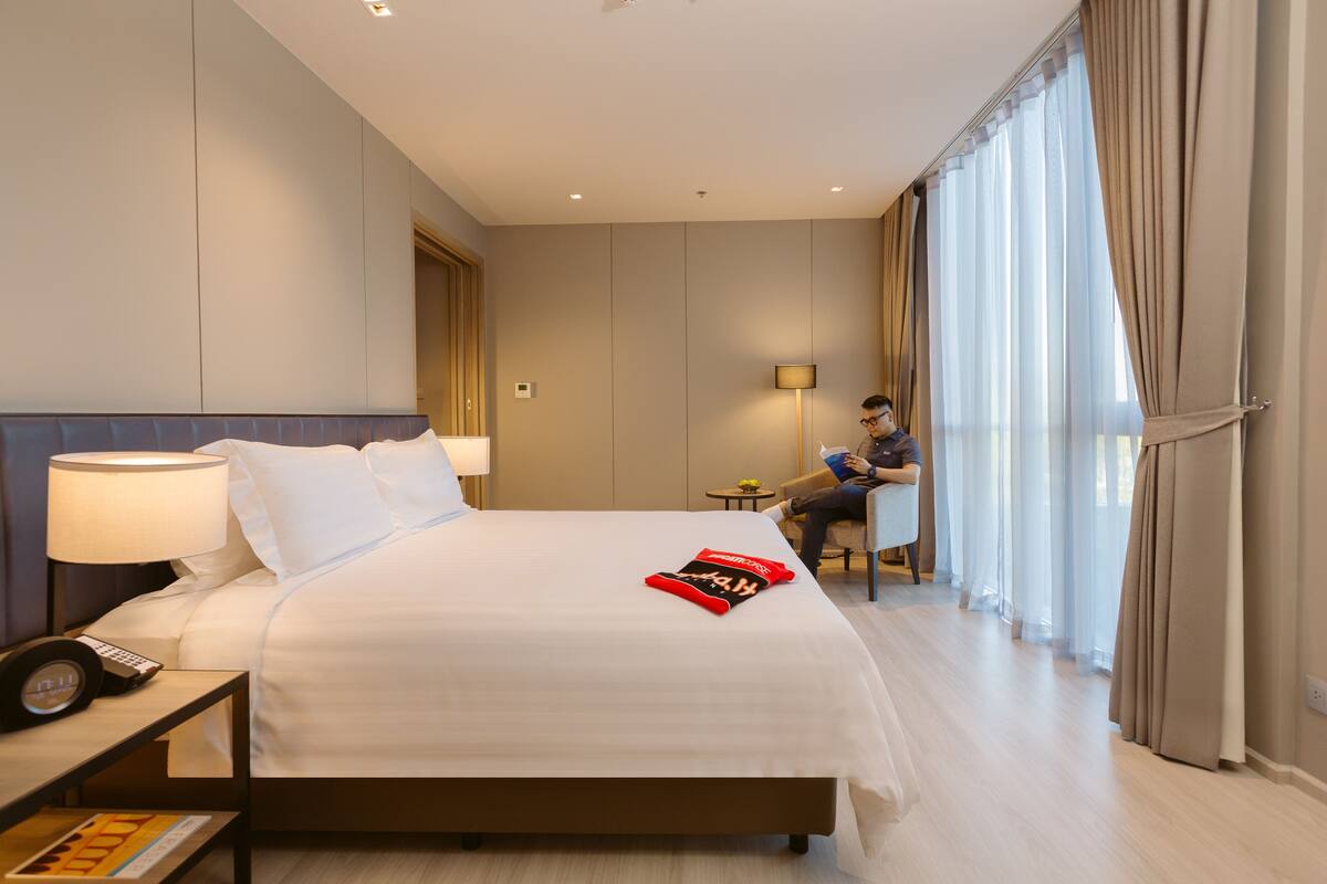 Ein Hotelzimmer mit einem großen Bett, einem Nachttisch mit Lampe, einem Sessel und einem Fenster mit Vorhängen.