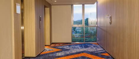 A hallway with a patterned carpet, wooden walls, and a large window offering a view of greenery and a building.