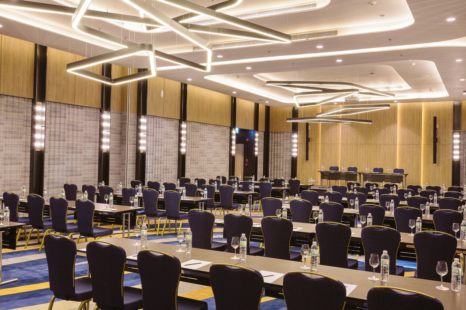 Sala de conferências com várias mesas e cadeiras, equipada com garrafas de água e copos, e luminárias modernas.