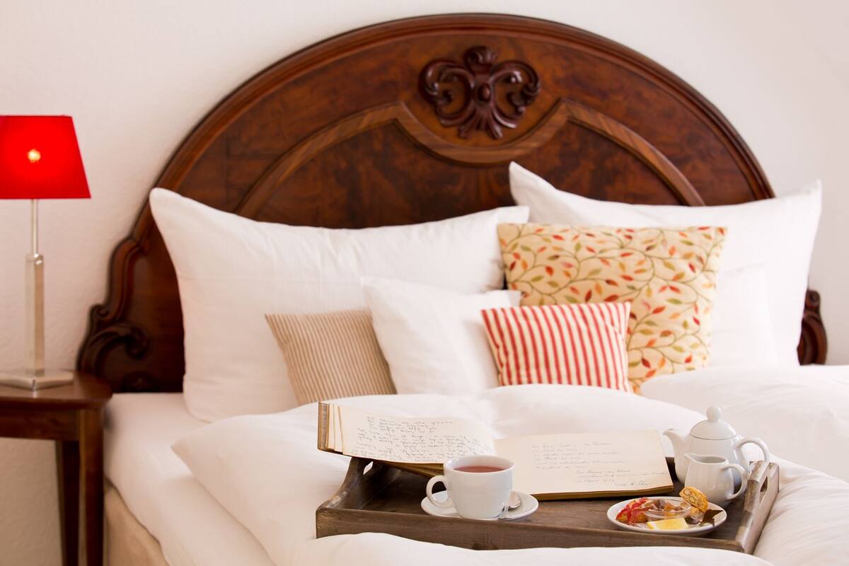 Un lit bien fait, avec une literie blanche et des coussins décoratifs, une table de chevet avec une lampe rouge, et un plateau contenant un livre, une tasse de thé et une assiette de nourriture.