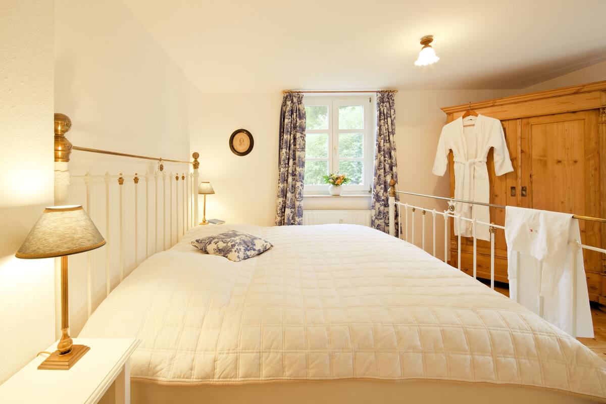 Une chambre à coucher comprenant un lit, une lampe de chevet, une fenêtre avec des rideaux, une armoire en bois et un porte-manteau mural.