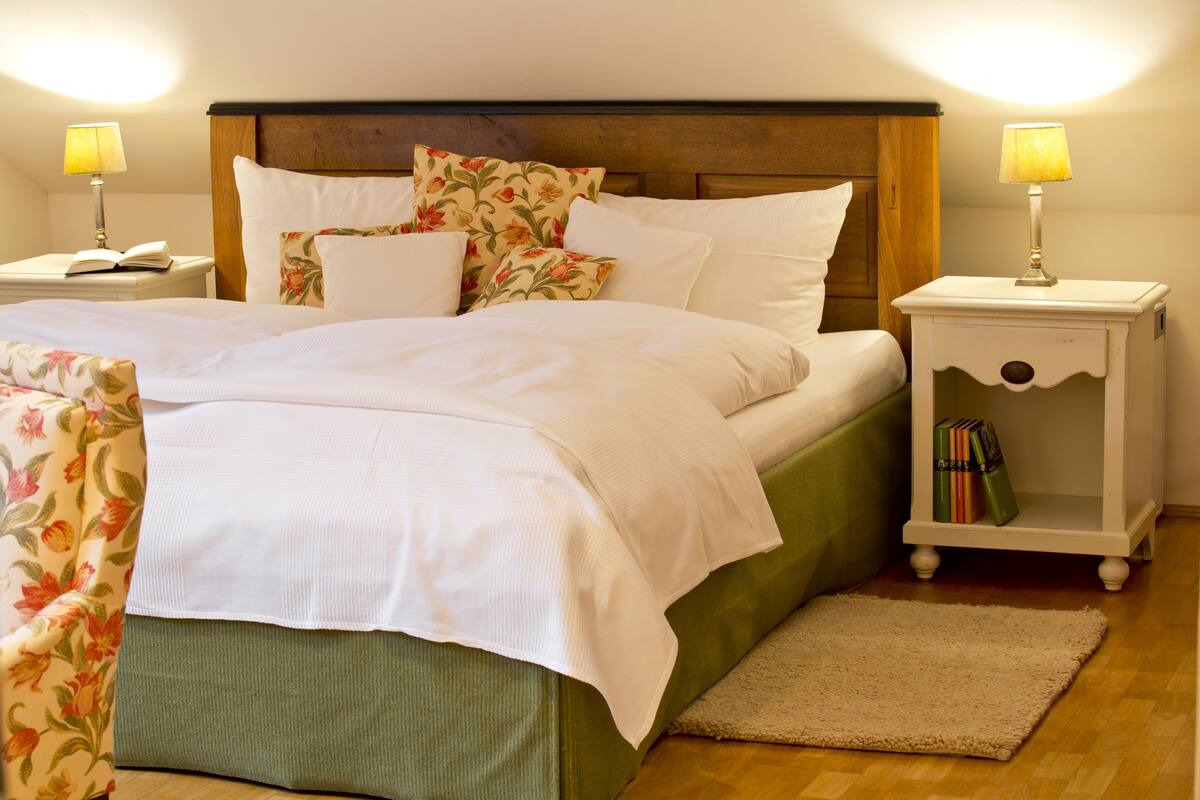 Un lit bien fait, avec du linge de lit blanc, une tête de lit recouverte d’un tissu vert et des coussins à motifs floraux. On y trouve deux tables de chevet avec des lampes et une petite étagère avec des livres.
