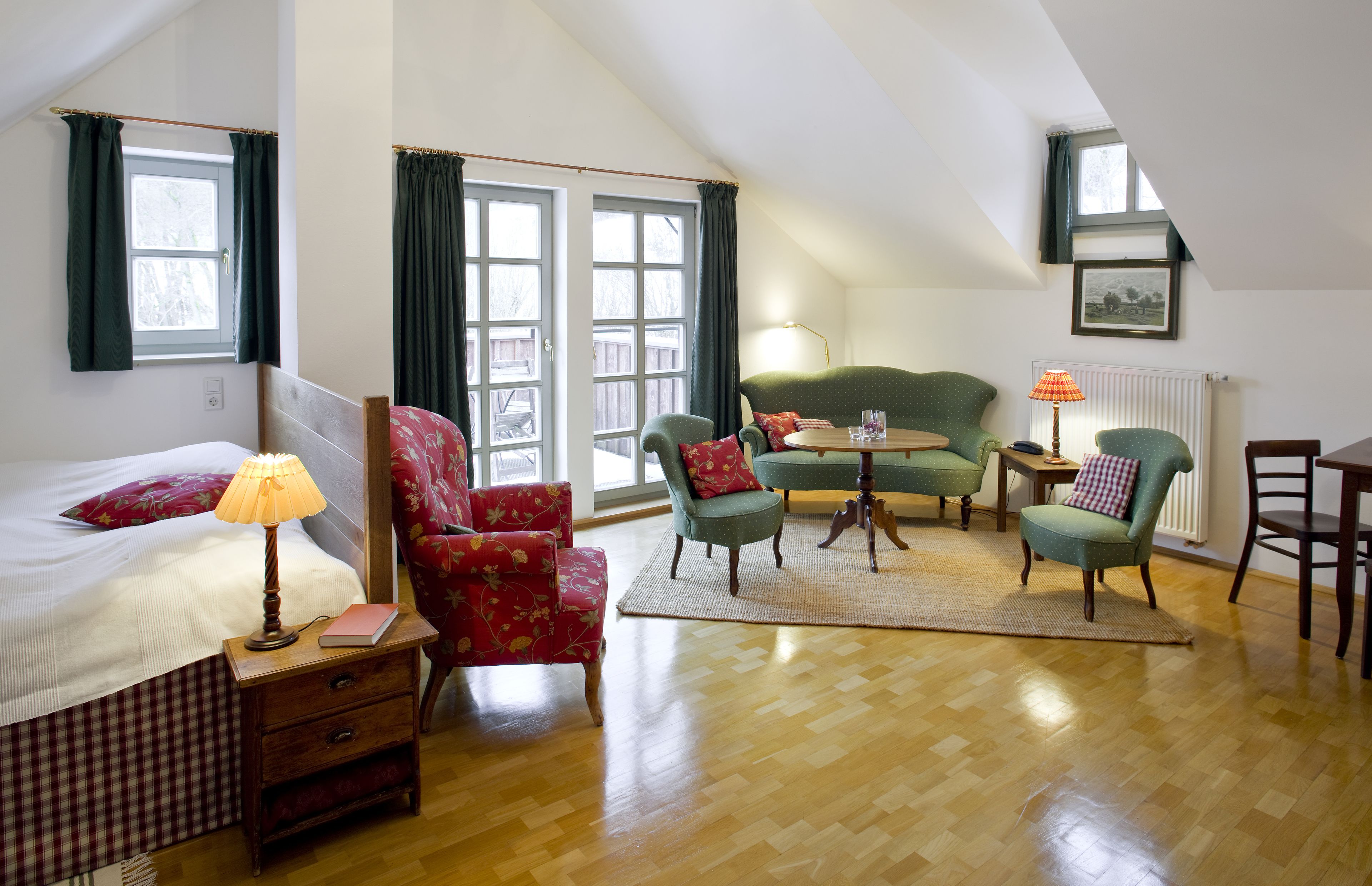 A cozy room with a bed, a desk, and two chairs. There is a window with curtains, a lamp, and a radiator.