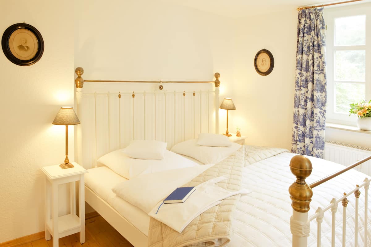 Un lit bien fait, avec du linge de lit blanc, deux tables de chevet avec des lampes et une fenêtre ornée d’un rideau à motifs floraux bleus.