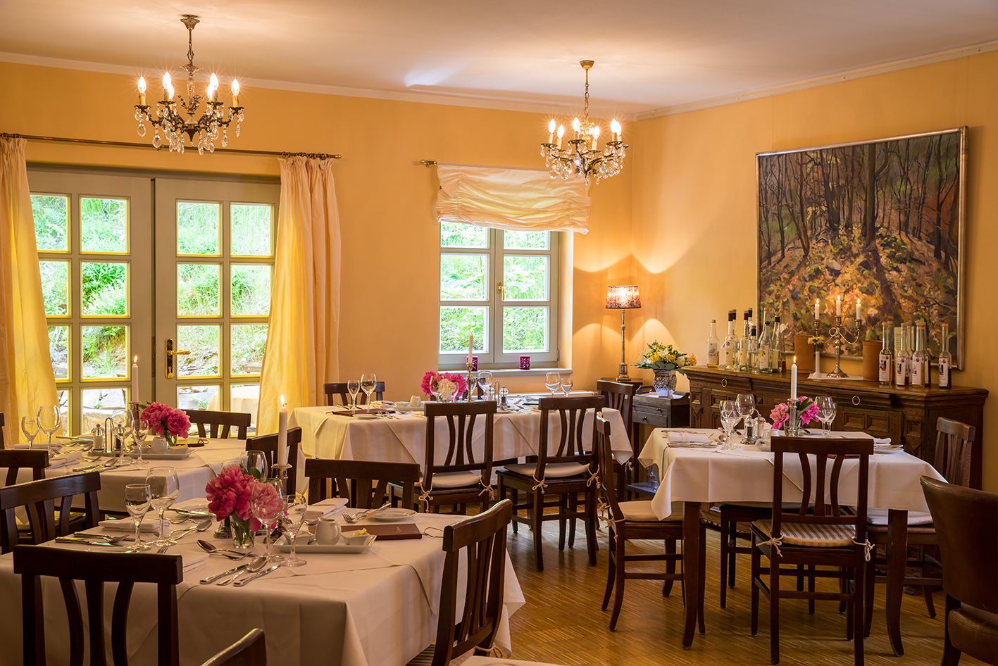 Une salle à manger avec plusieurs tables dressées, des lustres et un grand tableau accroché au mur.