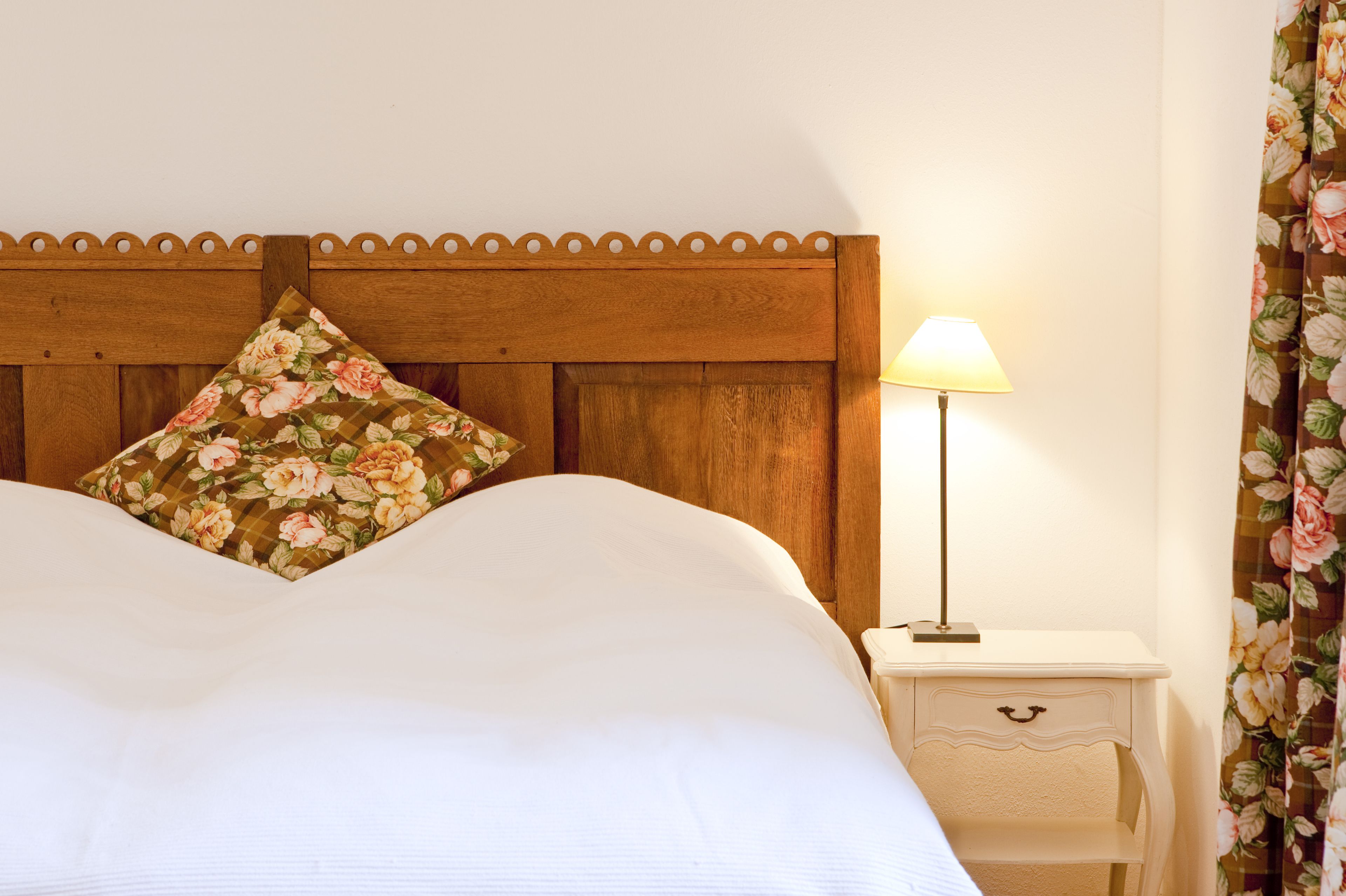 Una cama bien hecha con un cojín floral, un cabecero de madera, una lámpara de noche y una mesita de noche pequeña con un cajón.