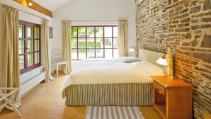 A bedroom with a large bed, a wooden nightstand, a stone wall, and a view of greenery outside. - Romantik Hotel Landhaus Bärenmühle (Frankenau)