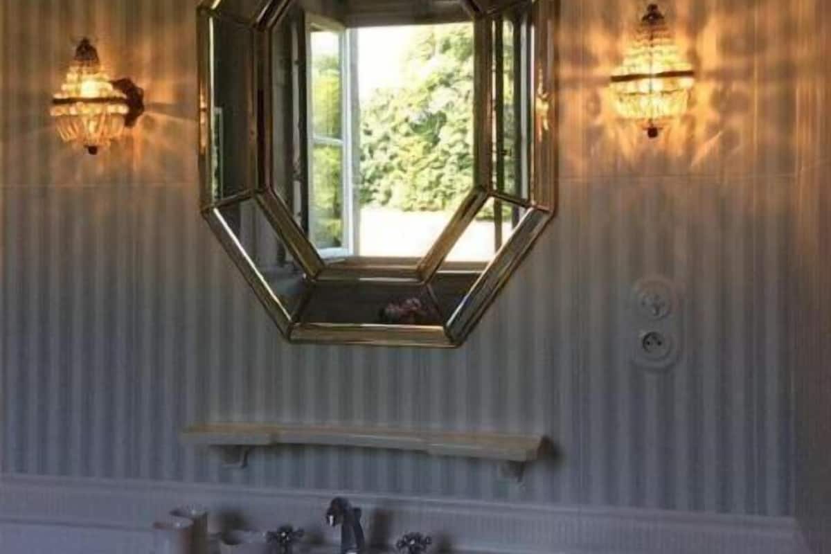 A bathroom with a large octagonal mirror, two wall-mounted sconces, a wooden vanity, and a window with a view of greenery.