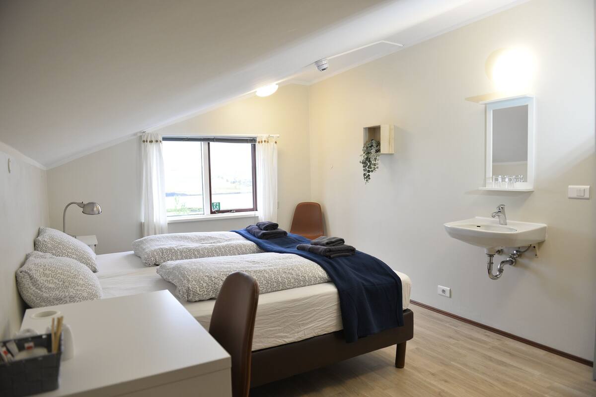 A bedroom with two beds, a desk, a chair, a window with curtains, a bedside lamp, a wall-mounted sink, and a mirror.