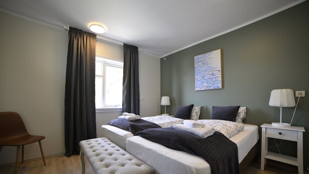 A bedroom with a bed, two bedside tables, a bench, a chair, and a window with curtains.