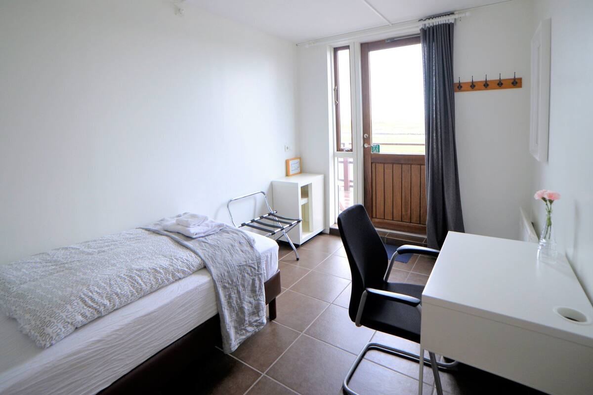 A small, clean room with a bed, a desk, and a chair. There is a window with curtains, a coat rack, and a small table with a picture frame.
