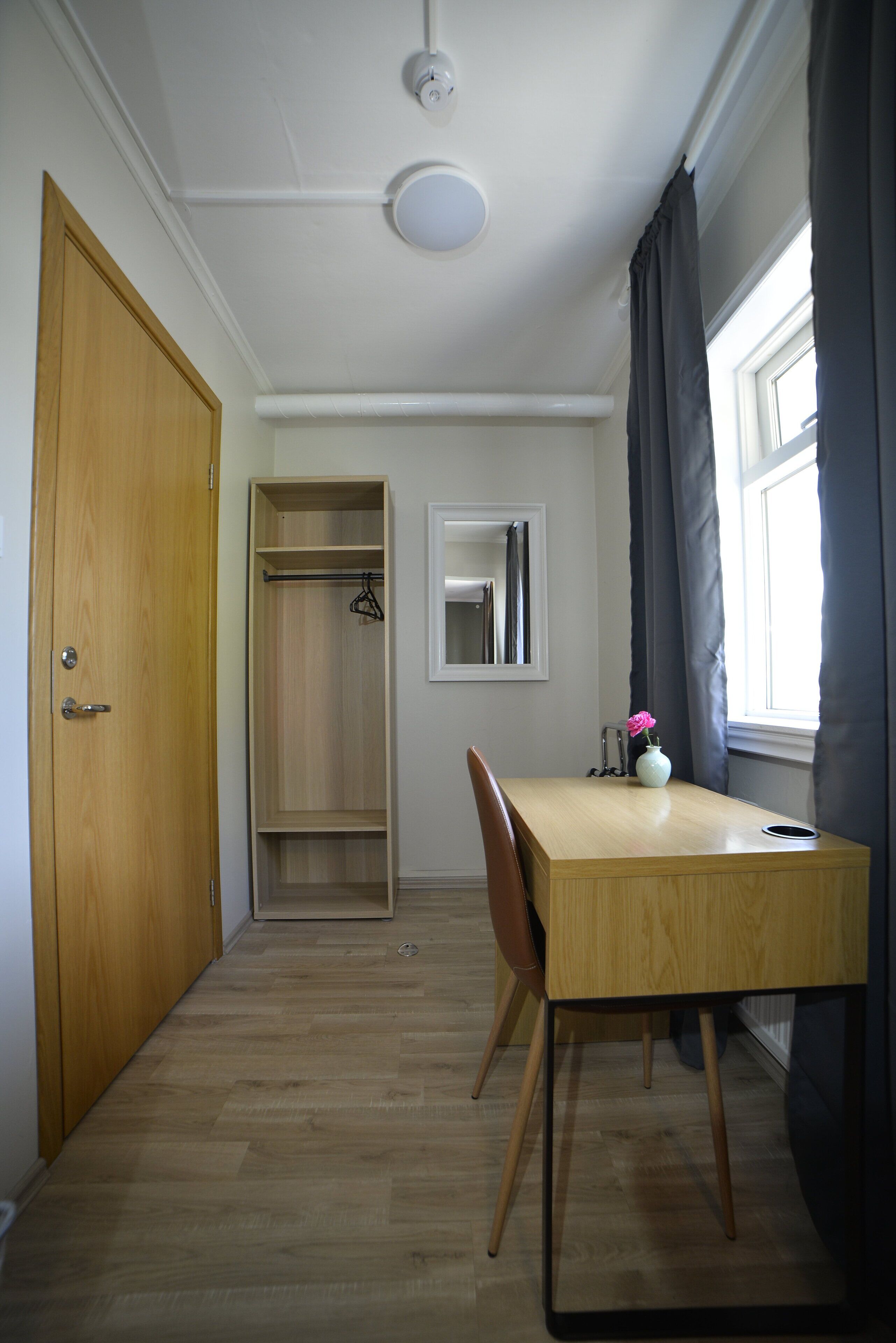 A room with a wooden door, a wooden table with a chair, a window with curtains, and a wardrobe.