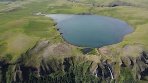 Une vue aérienne d’un lac entouré de collines verdoyantes et d’une zone boisée où se trouve une cascade.