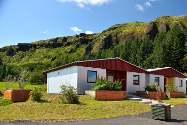 A modern building with a red roof and white walls, surrounded by greenery and mountains. - Klausturhof Guesthouse (Kirkjubaejarklaustur)