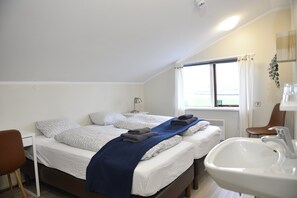 A bedroom with two beds, a desk, a chair, a window with curtains, and a bathroom with a sink. - Klausturhof Guesthouse (Kirkjubaejarklaustur)