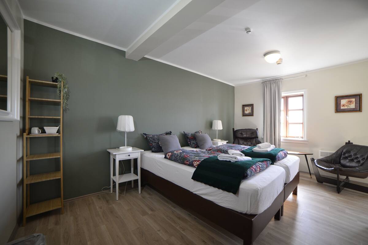 A bedroom with two beds, a wooden shelf, a desk, a chair, and a window with curtains.