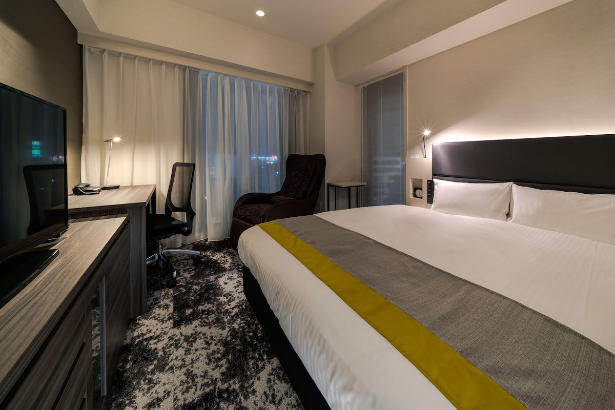 Ein Hotelzimmer mit einem großen Bett, einem Schreibtisch mit Computer, einem Sessel und einem Fernseher.