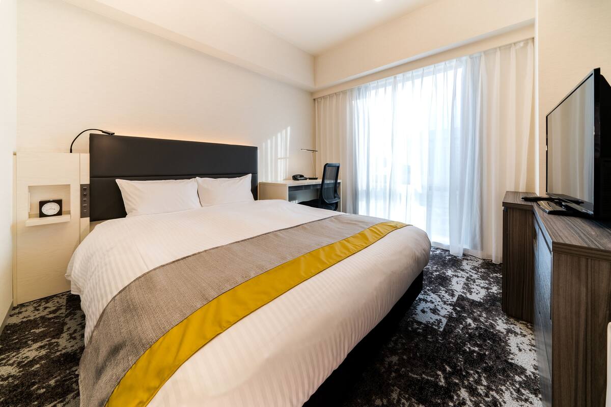 Ein Hotelzimmer mit einem großen Bett, einem Schreibtisch, einem Fernseher und einem Fenster mit Vorhängen.