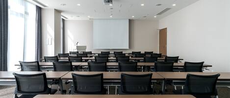 Ein Konferenzraum mit mehreren Tischen und Stühlen, einer Projektionsleinwand und einem großen Fenster mit Vorhängen.