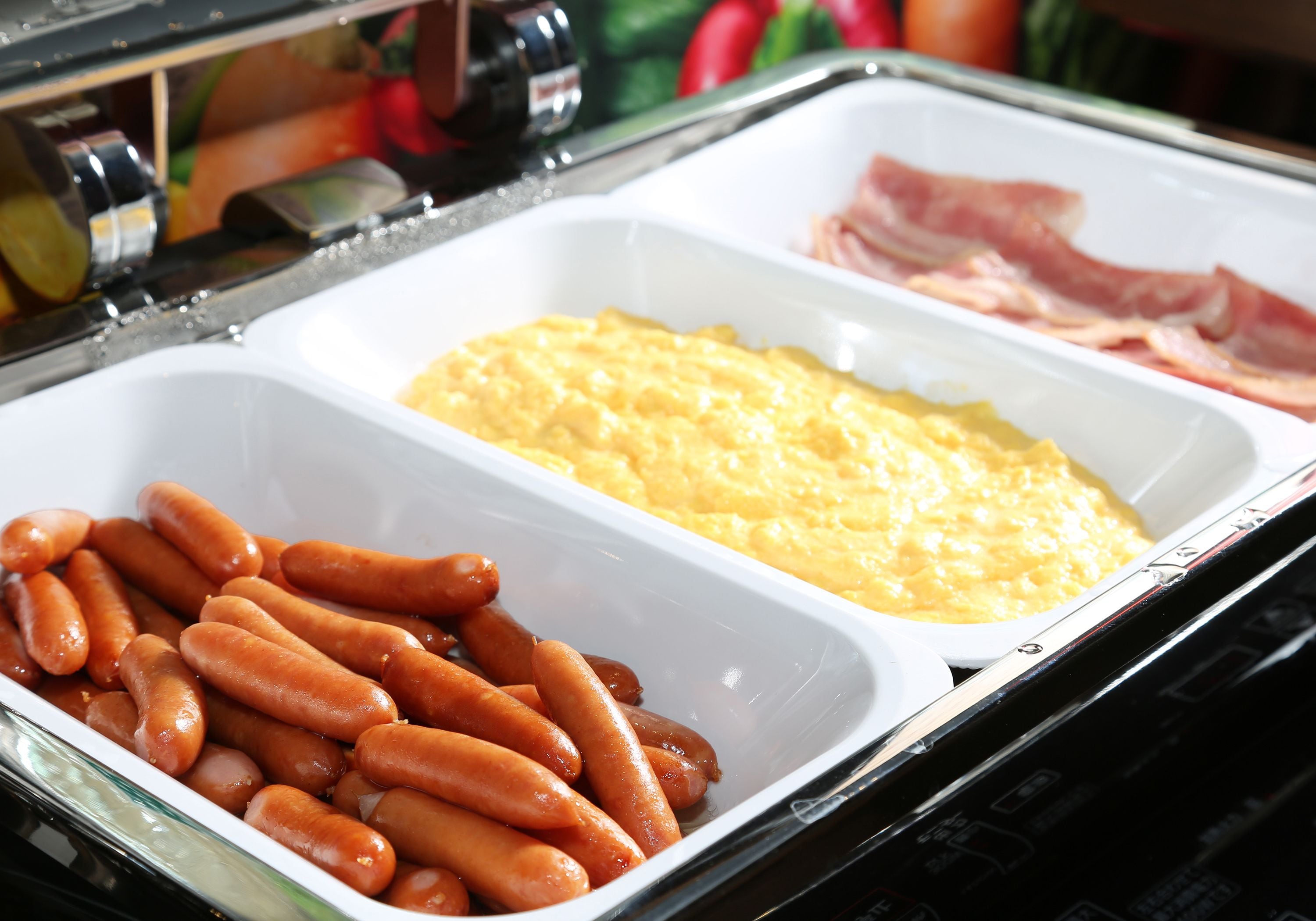 A buffet table with trays of sausages, scrambled eggs, and ham.