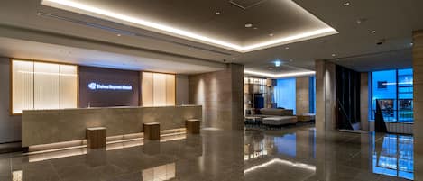 A modern hotel lobby with a reception desk, a seating area, and a view of the outside.