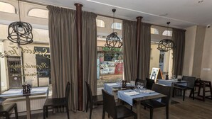 Un comedor con mesas dispuestas para dos, ventanas amplias con cortinas, y vista a la calle.