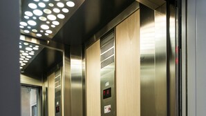 Interior de un ascensor con paredes metálicas, un panel de madera y un panel de control con botones y una pantalla digital.