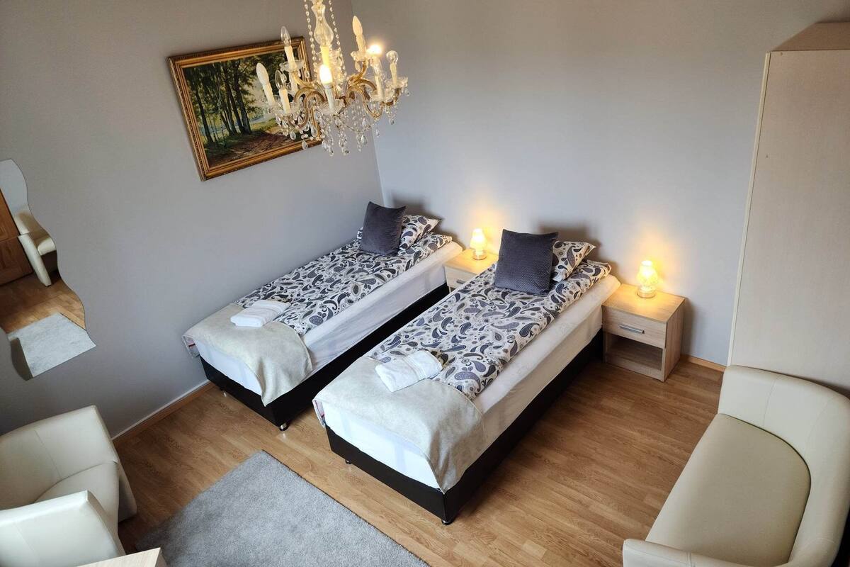 A bedroom with two beds, a chandelier, a painting, and a chair.