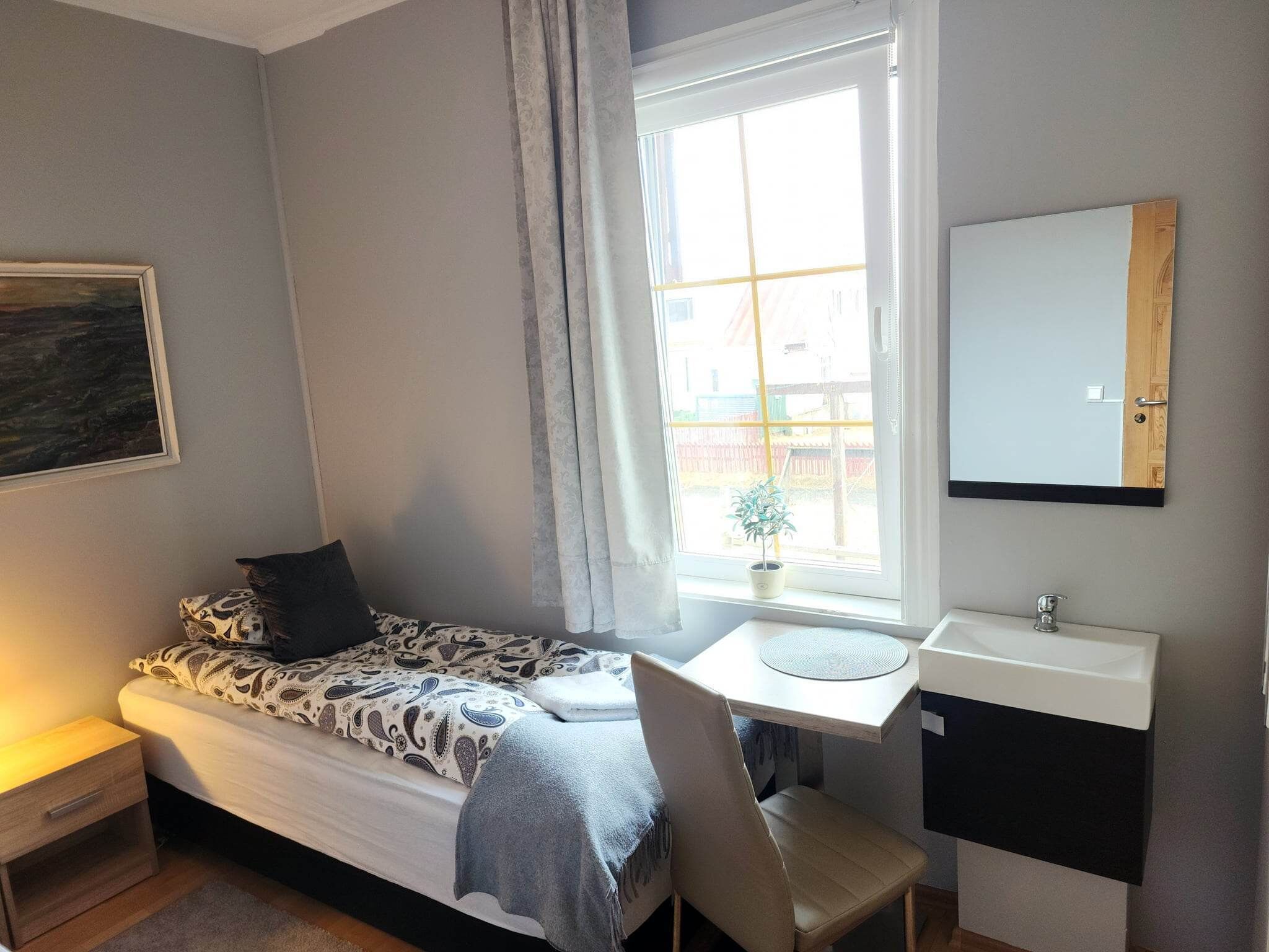 Um quarto pequeno e bem arrumado, com uma cama de solteiro, uma mesa de cabeceira, uma escrivaninha com cadeira, uma janela com cortinas, um vaso de planta e um espelho sobre a pia.